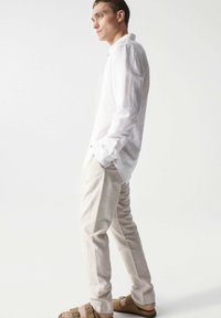 Camisa de manga larga de lino blanco, pantalones beige y sandalias marrones. La camisa cuenta con un cierre de botones, tiene un ajuste relajado y una textura ligera.