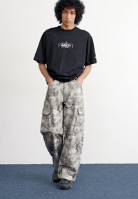 Camiseta negra de gran tamaño con estampado gráfico, combinada con pantalones cargo gris camuflaje que cuentan con múltiples bolsillos. El modelo lleva zapatillas verdes.
