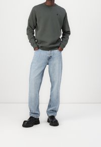 Felpa verde scuro con scollo rotondo, abbinata a jeans denim azzurri chiari a taglio largo e scarpe nere con suola spessa. Design semplice e stile casual.