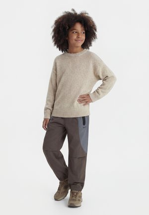 Otrok z kodrastimi lasmi v bež puloverju pletenine, temnih hlačah v barvnih blokih in rjavih supergah, stoječ z roko na boku.