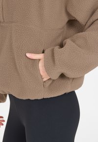 Brauner Fleece-Pullover mit strukturierter Oberfläche, Reißverschlusstasche und lockerer Passform. Getragen über schwarzen Leggings und mit weiten Ärmeln.