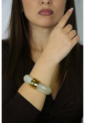 Brazalete blanco translúcido con detalles dorados, con un diseño suave y redondeado. Se lleva en la muñeca, destacando un contraste con el tono de piel.