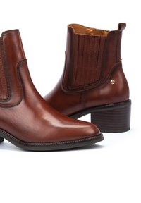 Botas de tobillo de cuero marrón con punta afilada, paneles laterales elásticos y un tacón apilado. Presentan detalles de costura y un acento dorado.