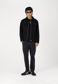 Casaco preto com fecho éclair e dois bolsos frontais, combinado com uma t-shirt escura e calças texturizadas escuras. Ténis pretos com riscas brancas.