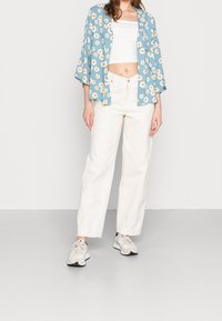 Kimono floreale azzurro chiaro con margherite bianche, abbinato a jeans a gamba larga di un bianco sporco e sneaker beige. Tessuto morbido, vestibilità rilassata.