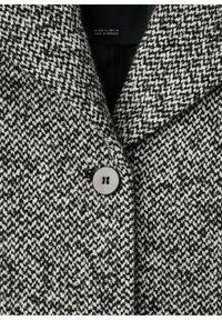 Cappotto in lana testurizzata bianco e nero con colletto a intaglio e chiusura a bottone rotondo sul davanti.