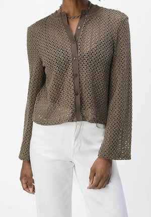 Cardigan marron à manches longues en crochet avec boutons porté sur un sous-vêtement marron, associé à un pantalon blanc, montré des épaules aux hanches.