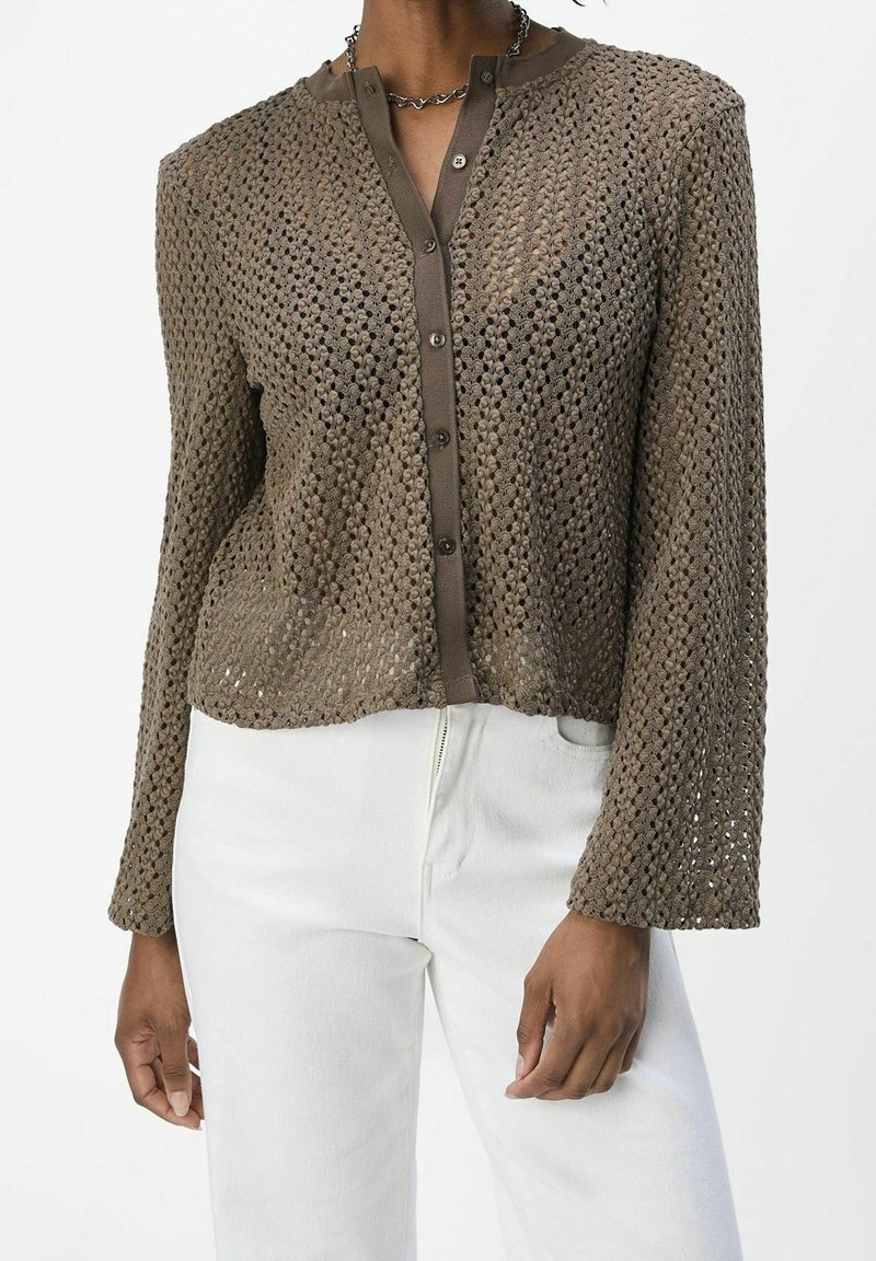 Cardigan marron à manches longues en crochet avec boutons porté sur un sous-vêtement marron, associé à un pantalon blanc, montré des épaules aux hanches.