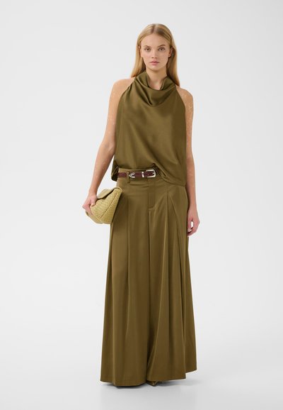 Femme debout portant un haut sans manches vert olive avec un pantalon à jambes larges, tenant une pochette matelassée beige.