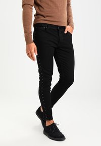 Jeans skinny de mezclilla negra con detalles de cordones en los laterales, tejido texturizado y bolsillos minimalistas, acompañados de zapatos de ante negros.