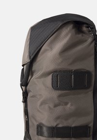 Un sac à dos gris foncé avec une texture noire accentuée, un ajusteur de sangle latéral et un rabat supérieur avec un motif en grille. Des coutures renforcées sont visibles.