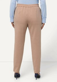Pantaloni tapered marrone chiaro con vita elasticizzata, tessuto liscio e vestibilità sartoriale. Abbinamento con tacchi blu scuri visibile.