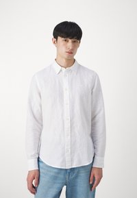 Camicia bianca di lino con colletto, maniche lunghe e orlo arrotondato. Presenta un delicato ricamo sul petto e bottoni chiari.