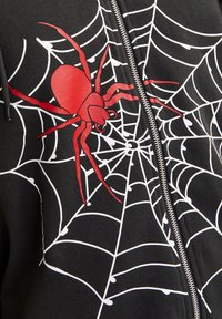 Sudadera negra con cremallera que presenta una gran araña roja y un diseño de telaraña blanca. El material parece suave y texturizado, adecuado para el uso diario.