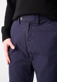 Námornícke modré bavlnené chinos s hladkým predným dielom, zapínaním na gombík a bočnými vreckami. Hladká textúra a strih s rovným nohavicami, s jemnými detailmi šitia.