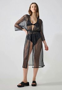 Kimono trasparente nero con maniche a tre quarti, vita con cordoncino e un orlo leggermente più lungo. Indossato sopra un bikini nero.