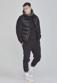 Juodas paminkštintas liemenė virš juodos hoodie, derinama su juodomis sportinėmis kelnėmis ir baltomis aukštakulnėmis sportinėmis batelėmis. Yra logotipų detalės ir raukšlių rankogaliai.