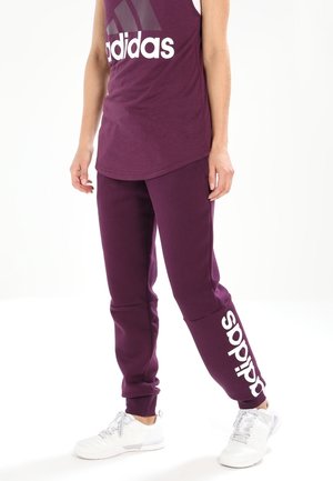 Persona que lleva pantalones deportivos morados de Adidas y una camiseta sin mangas a juego con el logo blanco de Adidas y zapatillas deportivas blancas.