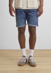 Shorts di denim con risvolti, colore blu scuro, abbinati a calze bianche alla caviglia e scarpe da ginnastica in suede marrone con strisce bianche.