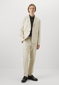 Beige kavaj med knappslag och framfickor, kombinerad med en svart prickig skjorta och beige byxor; komplett med svarta skor.