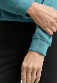 Morska bluza z okrągłym dekoltem i ściągaczami przy rękawach, zestawiona z czarnym materiałem, prezentująca gładką fakturę i swobodny design.