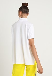 Camisa de polo de manga corta blanca con un corte relajado, que presenta un cuello alto y un tejido suave y texturizado. Llevada con pantalones amarillos.