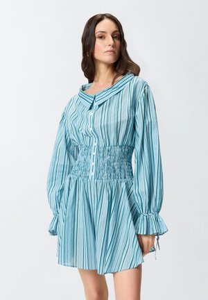 Femme portant une robe à rayures verticales bleu et blanc avec des manches longues bouffantes, une taille smockée et un devant boutonné, debout devant un fond uni.