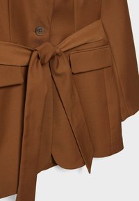 Blazer marron avec ceinture en tissu nouée, poches à rabat et une seule fermeture à bouton noir à l'avant, présenté sur un fond uni.