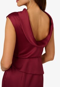 Burgundské saténové šaty s draped zády, bez rukávů a vrstveným peplum detailem. Lesklý povrch zvýrazňuje hladkou texturu.