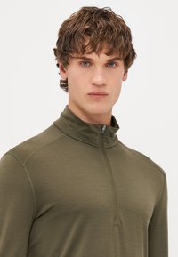 Olivovo zelený zip-up pulover vyrobený z mäkkého a elastického materiálu. Obsahuje vysoký golier a jemné švy, ktoré vytvárajú priliehavý vzhľad.