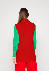 Gilet rouge en tricot avec un col montant, porté sur un chemisier vert à manches longues avec des détails de boutons aux poignets, associé à un pantalon rouge.