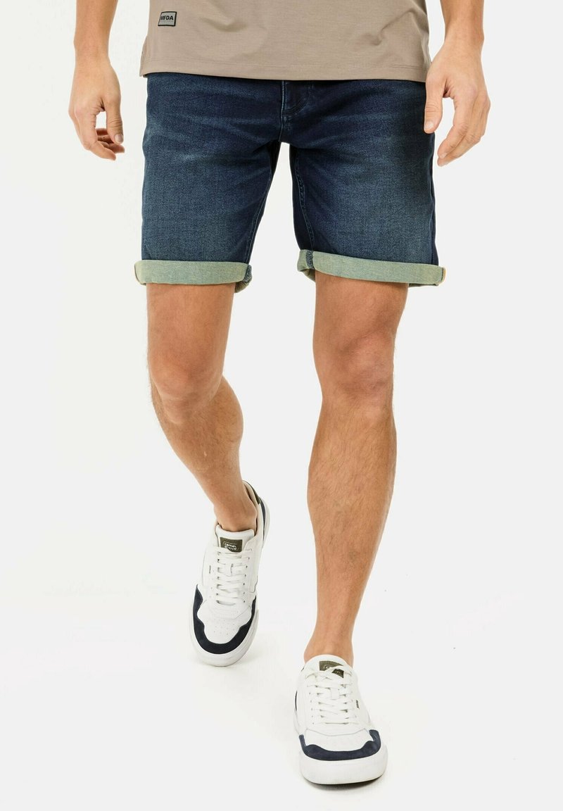 Homme marchant portant un short en jean foncé retroussé, une chemise beige et des baskets blanches avec des détails noirs sur fond blanc.