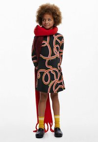 Schwarzes Kleid mit bunten, verschlungenen Mustern und gelbem Text, kombiniert mit einem langen, roten Schal. Gelbe Socken und schwarze Schuhe vervollständigen das Outfit.