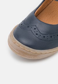 Zapato de piel de color marino con detalles de brogue decorativos, punta redonda y suela de goma beige flexible. Interior texturizado con costuras visibles.