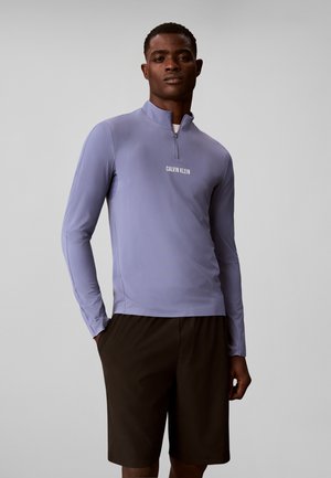 Мъж, носещ лилава блуза с дълги ръкави Calvin Klein с цип и черни шорти, стои с една ръка в джоба пред сив фон.