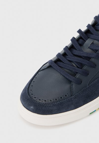 Marinblå sneaker med en slät lädertå och texturerad mockaöverkjol, med matchande snören och en vit gummisula med färgglada detaljer.