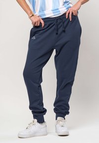 Osoba ubrana w granatowe dresy z ściągaczem w talii, białe buty sportowe oraz niebiesko-białą paskowaną koszulę podnoszącą jej brzeg.