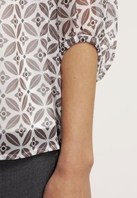 Transparente Bluse mit einem sich wiederholenden geometrischen Muster in Grau und Weiß, mit gerafften Bündchen und lockerer Passform, kombiniert mit strukturiertem grauem Stoff.
