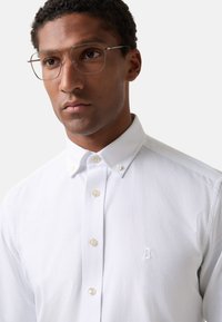 Witte button-up shirt met een kraag en geborduurde letter "B" op de borst, gemaakt van een gladde stof met een subtiele glans.