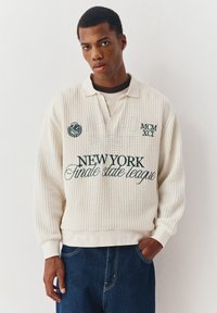 Jonge man met een gebreide crèmekleurige trui met de tekst "NEW YORK finale state league" en donkerblauwe spijkerbroek, staand tegen een effen witte muur.