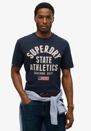 Mørkeblå bomulds T-shirt med fed cremefarvet tekst, der står "SUPERDRY STATE ATHLETICS." Indeholder et amerikansk flaggrafik og en rund halsudskæring.