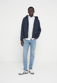 Μπλε μπουφάν με μόνωση και φερμουάρ, φορεμένο πάνω από ένα λευκό πουκάμισο με σχέδιο, συνδυασμένο με ανοιχτό μπλε τζιν και ασπρόμαυρα παπούτσια.