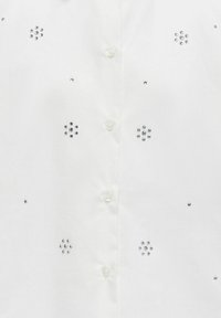 Camicia bianca con diverse decorazioni in strass disposte in motivi floreali e chiusura a bottoni sul davanti.