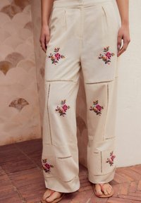 Pantaloni beige a gamba larga con ricami floreali, caratterizzati da dettagli a taglio e pieghe sul davanti, realizzati in morbido tessuto di cotone.