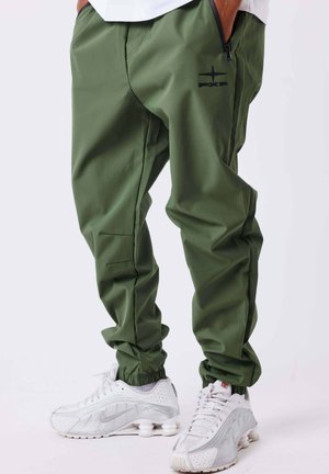 Personne portant un pantalon cargo vert olive et des baskets blanches Nike, debout les mains dans les poches devant un fond uni.