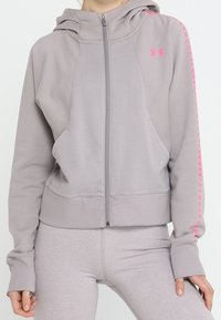 Persona che indossa una felpa grigia chiara con zip di Under Armour, con logo e testo rosa sulle maniche, abbinata a leggings coordinati.