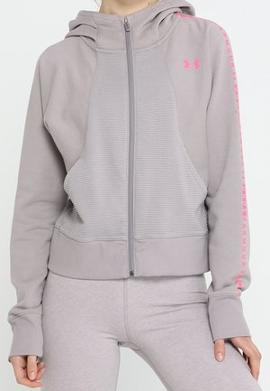 Personne portant un hoodie zippé gris clair Under Armour avec un logo rose et du texte sur les manches, associé à des leggings assortis.