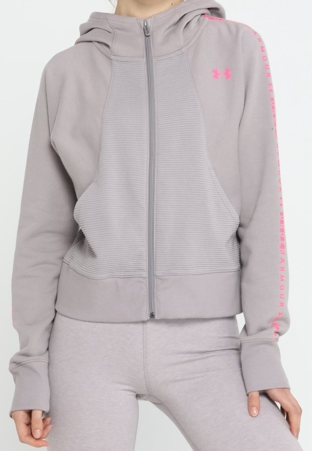 Persona che indossa una felpa grigia chiara con zip di Under Armour, con logo e testo rosa sulle maniche, abbinata a leggings coordinati.