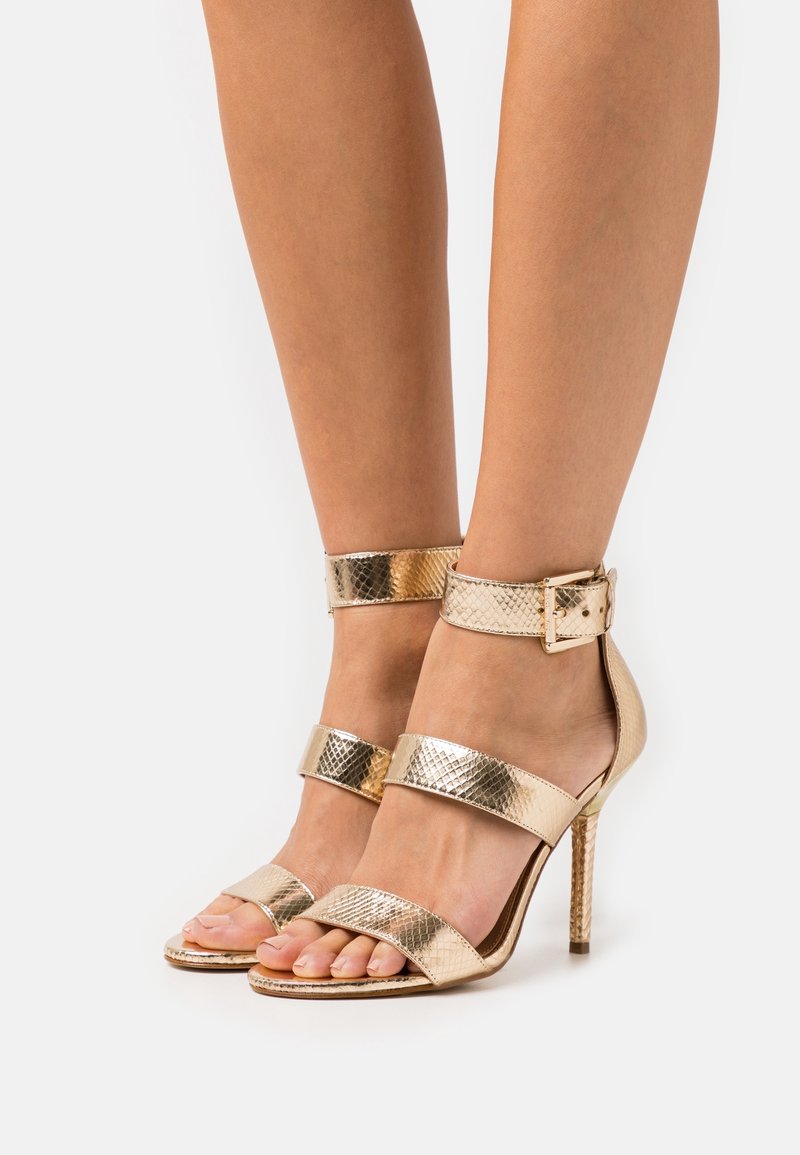 Jambes portant des sandales à talons hauts dorées avec trois brides texturées, y compris une bride de cheville avec boucle.