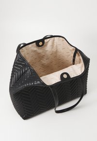 Sac à main noir matelassé avec un motif chevrons, doté d'un intérieur crème avec une marque répétée, de deux poignées et d'une fermeture à pression dorée.